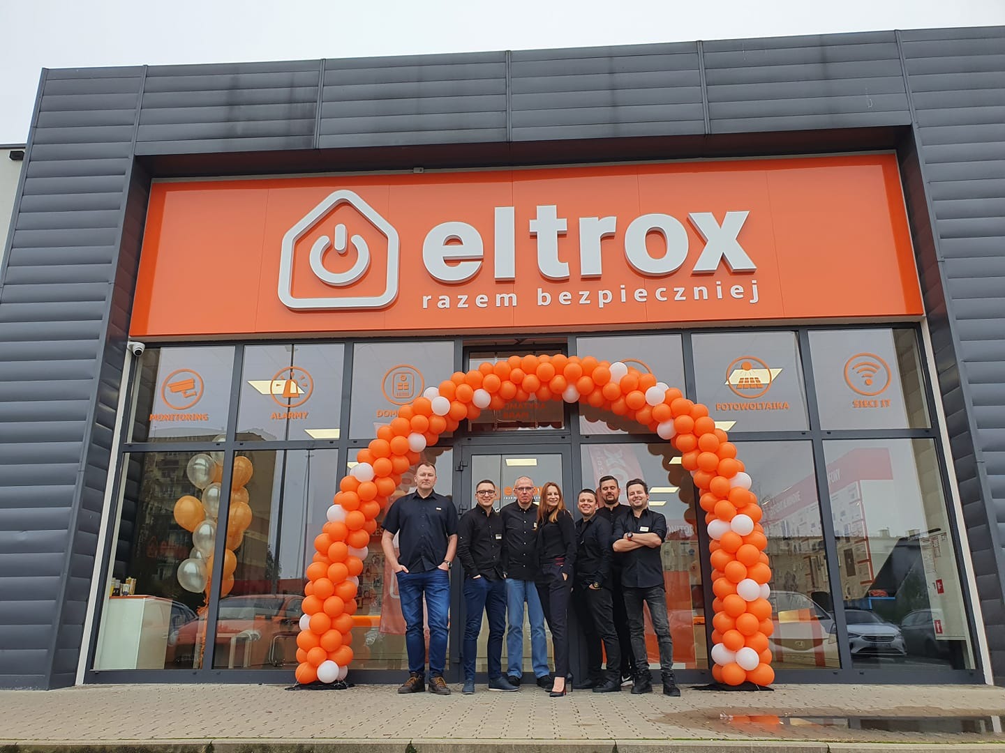 Grupa pracowników firmy Eltrox pozuje przed wejściem do sklepu z pomarańczowym szyldem i balonową girlandą. Na szybach widoczne grafiki z monitoringiem, alarmami, fotowoltaiką i siecią TV.