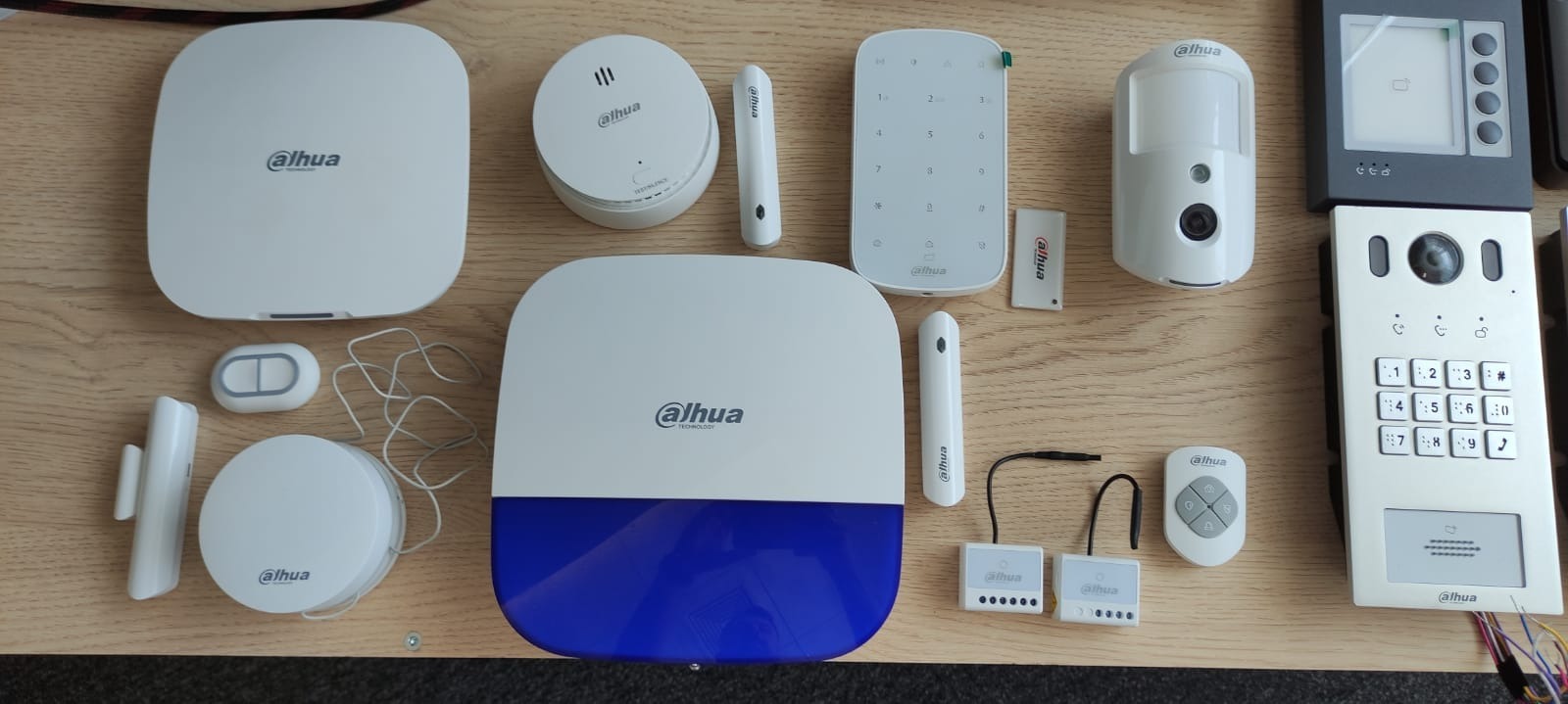 Zestaw komponentów systemu alarmowego Dahua ułożony na drewnianej powierzchni, zawierający centralę alarmową, czujniki ruchu, kontaktrony, klawiaturę numeryczną, manipulator, pilot, sygnalizator...