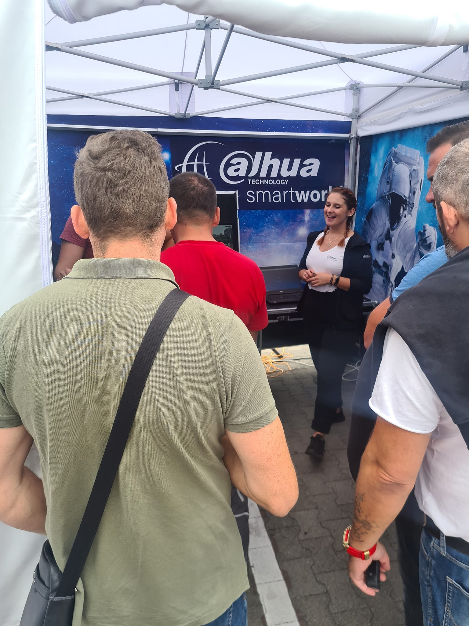 Prezentacja firmy Dahua na stoisku targowym, z uśmiechniętą kobietą w centrum, otoczoną przez potencjalnych klientów.