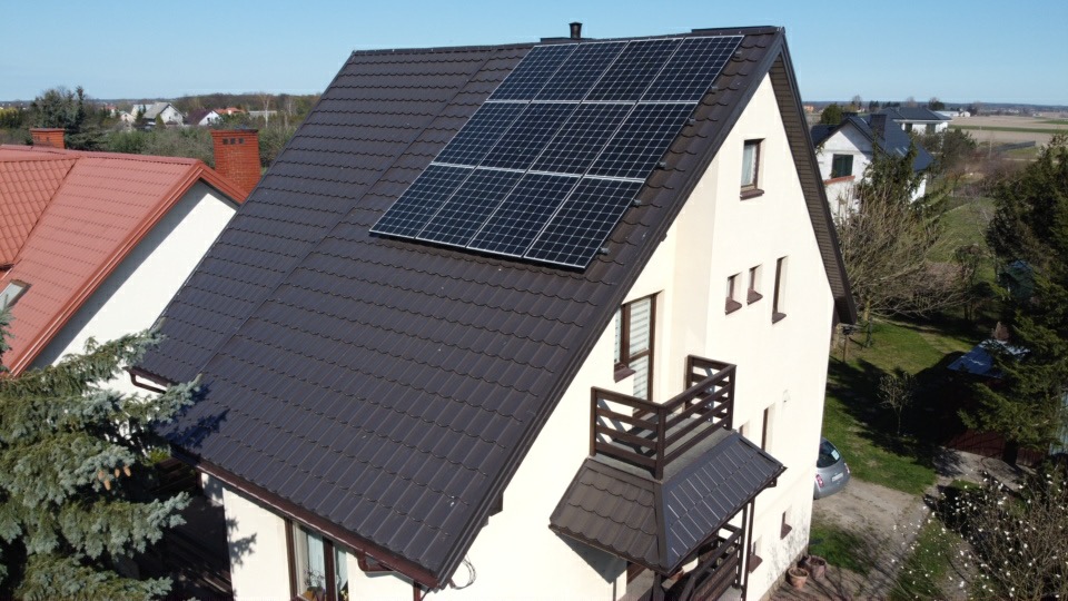 Dom jednorodzinny z ciemnobrązowym dachem, na którym zamontowane są panele fotowoltaiczne; widok z góry ukazuje również mały balkon i otoczenie domu.