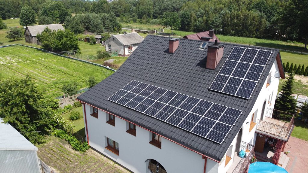 Widok z góry na dom jednorodzinny z panelami fotowoltaicznymi na dachu z dachówki ceramicznej, kominami i anteną satelitarną, otoczony zielenią.