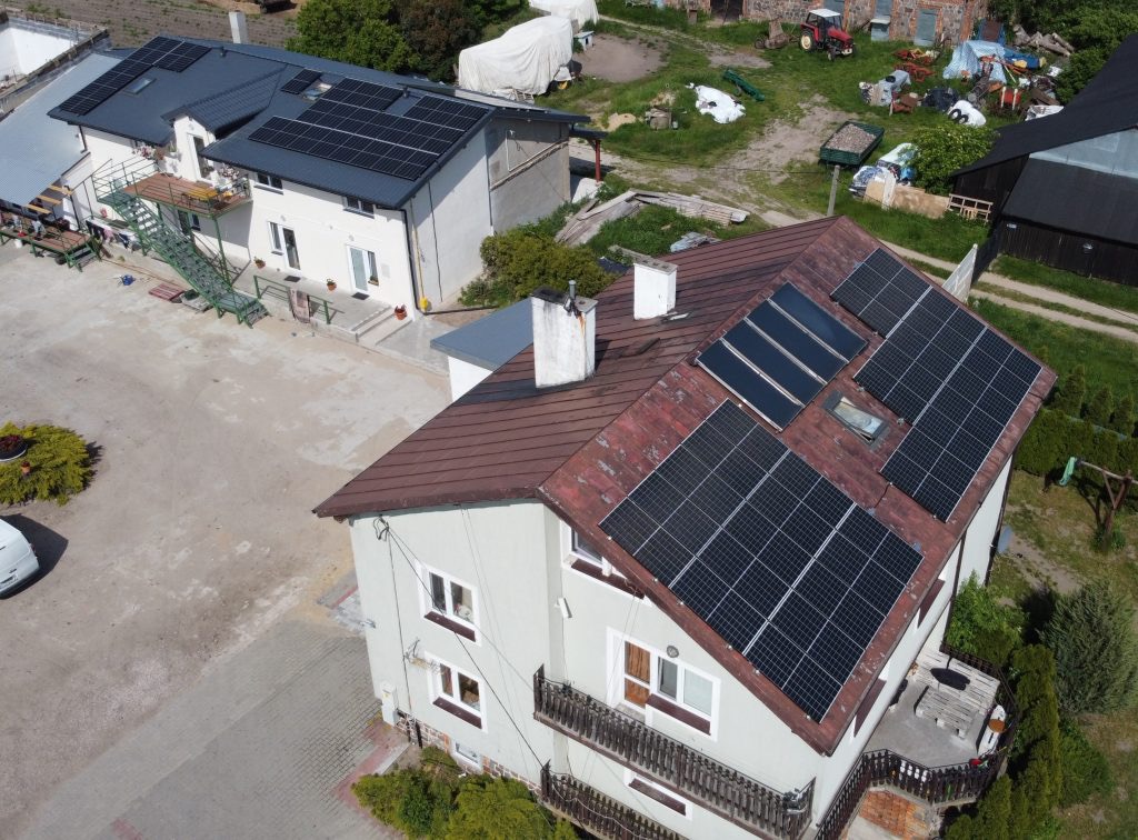 Widok z góry na dom z panelami słonecznymi na dachu, z widocznymi innymi budynkami i terenem zielonym w tle.