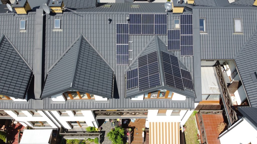 Widok z góry na dach szeregowca z szarej dachówki z zainstalowanymi panelami fotowoltaicznymi, widoczne okna dachowe i anteny satelitarne.