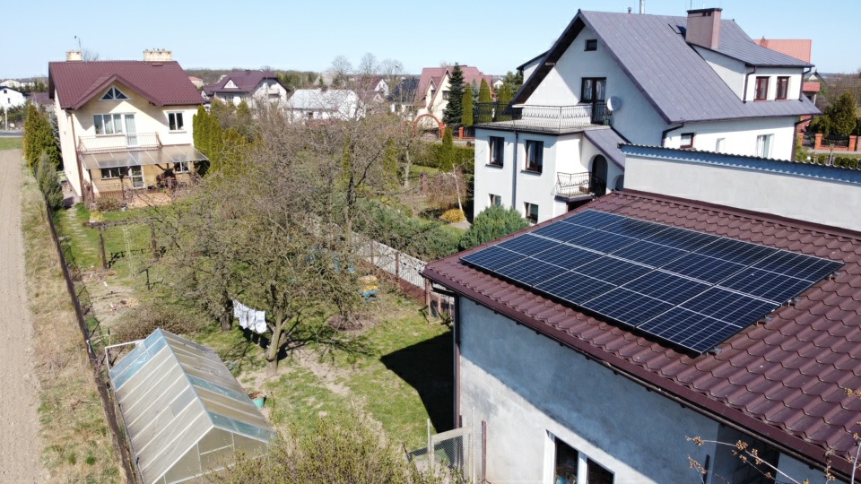 Panele fotowoltaiczne zamontowane na dachu domu jednorodzinnego z widokiem na ogród z szklarnią i suszącym się praniem na sznurku.