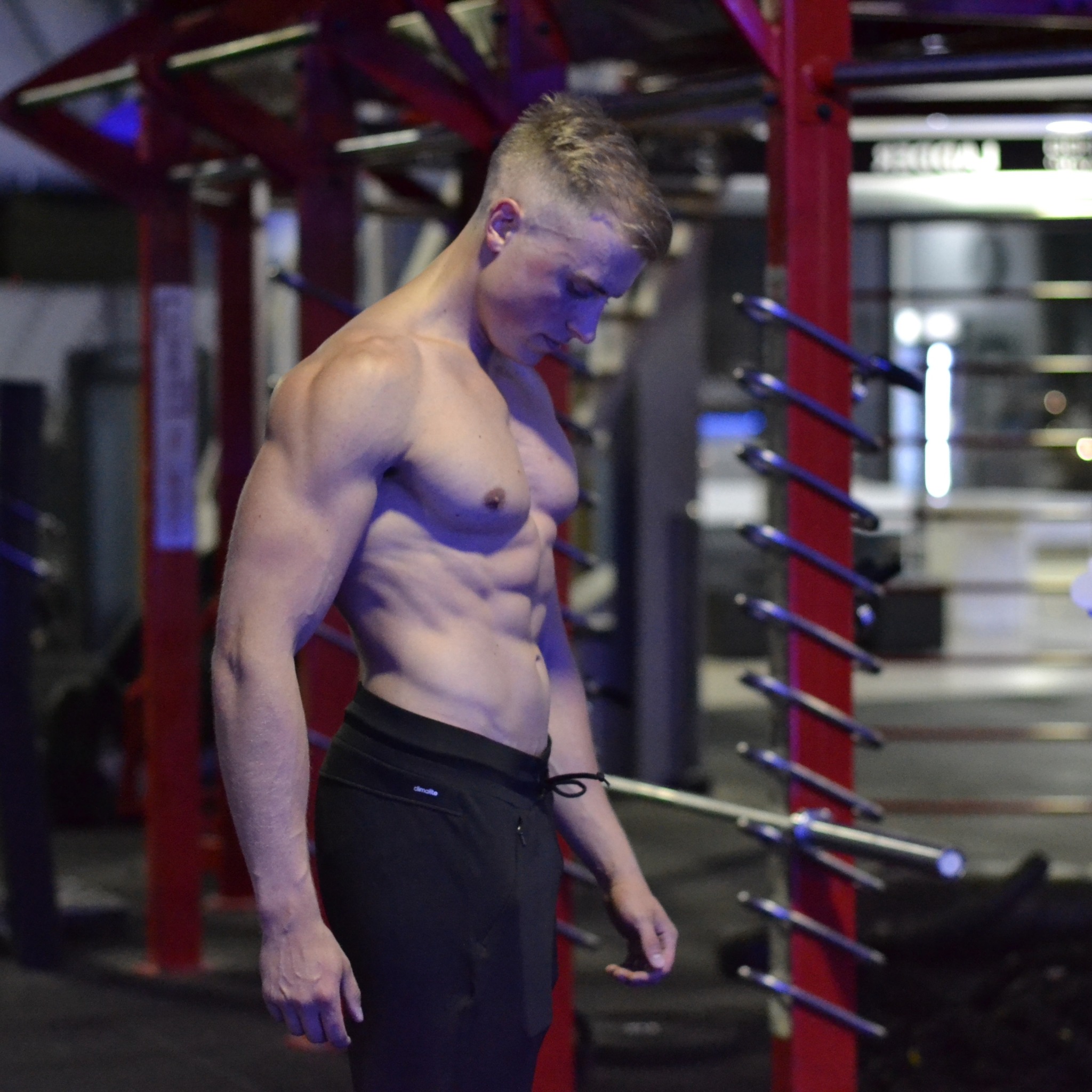 Mężczyzna z krótkimi blond włosami, bez koszulki, prezentuje wyrzeźbioną sylwetkę w siłowni, z widocznymi mięśniami brzucha i ramion, na tle czerwonej konstrukcji do ćwiczeń.