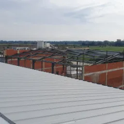 Konstrukcja stalowa hali w trakcie budowy, widok z dachu, z elementami murowanymi, na tle zieleni i zabudowań przemysłowych.