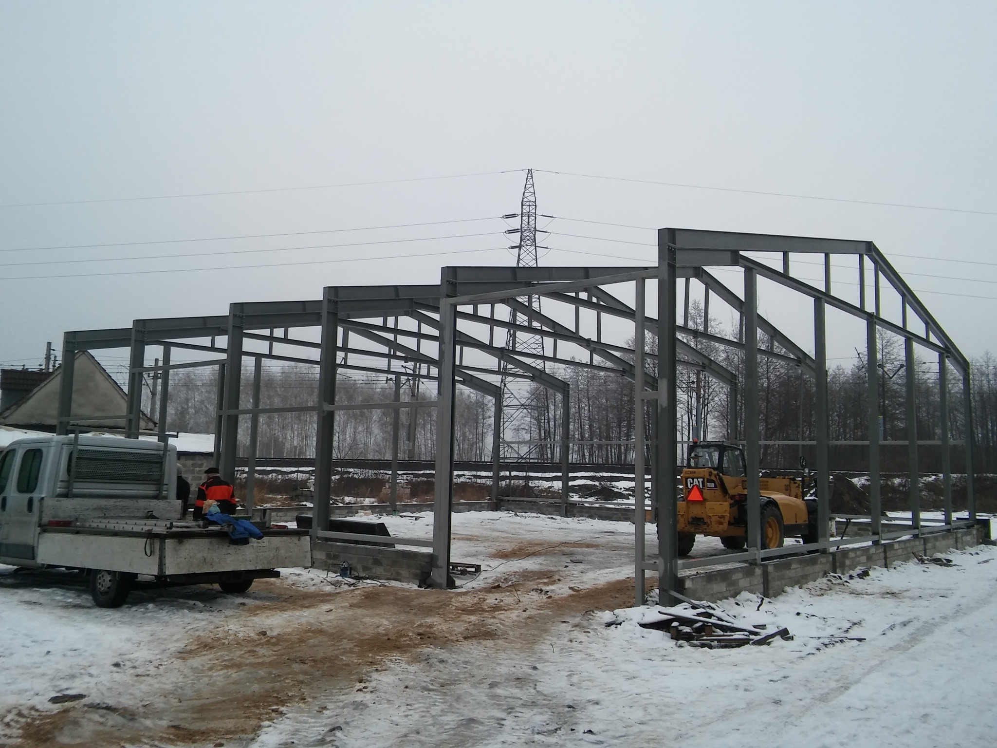 Stalowa konstrukcja hali w trakcie budowy, widoczny pracownik na pace dostawczego auta i maszyna budowlana CAT w tle, zimowa sceneria.