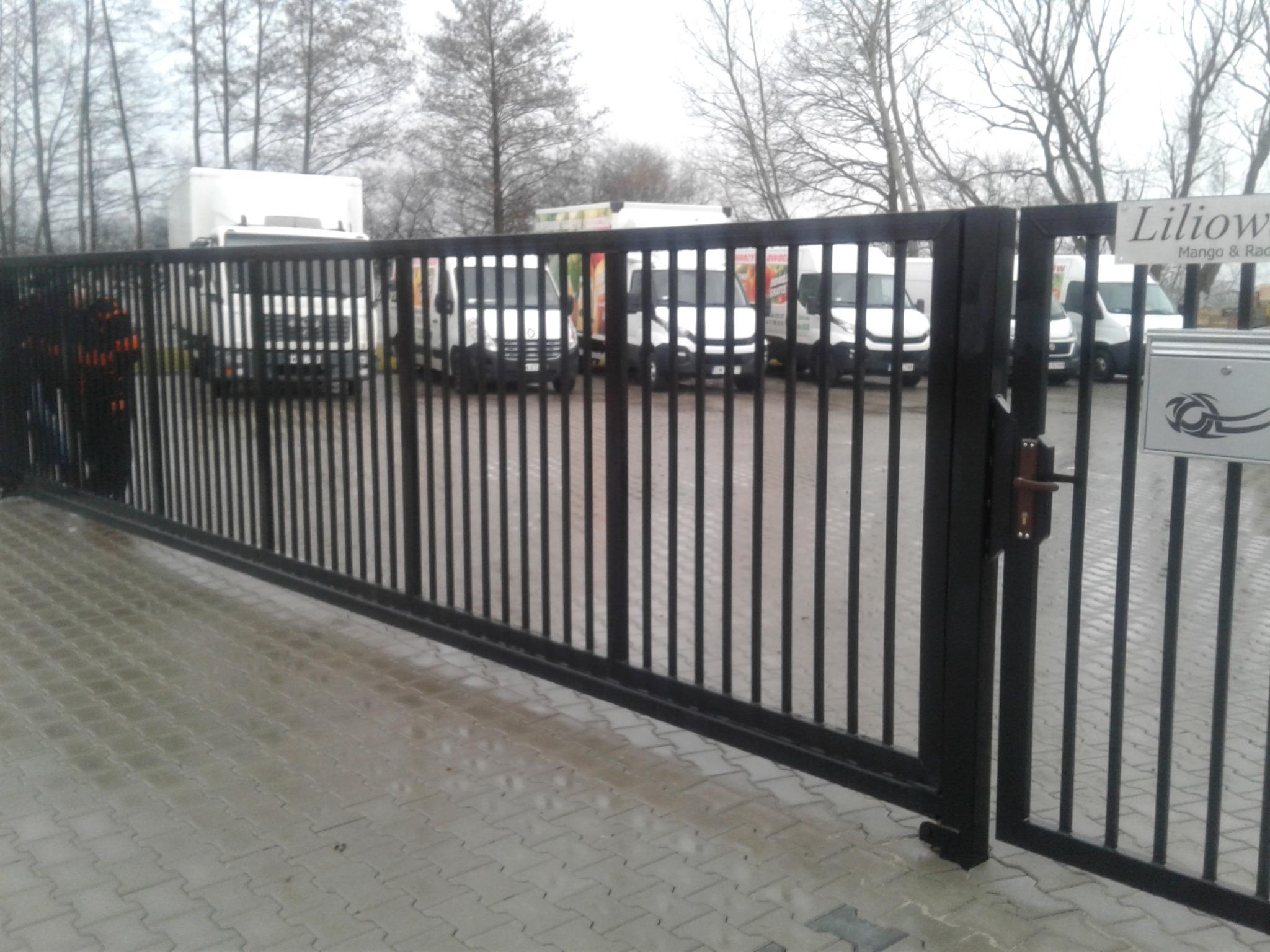 Czarna, metalowa brama przesuwna z pionowymi prętami, z widocznymi samochodami dostawczymi w tle i tabliczką z nazwą firmy.