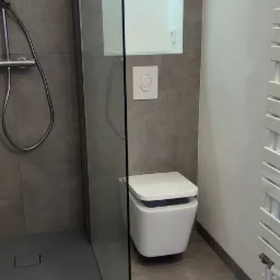 Nowoczesna łazienka z kabiną prysznicową typu walk-in, podwieszaną toaletą z przyciskiem w kształcie uśmiechu i grzejnikiem drabinkowym w kolorze białym, szare płytki na ścianach i podłodze.