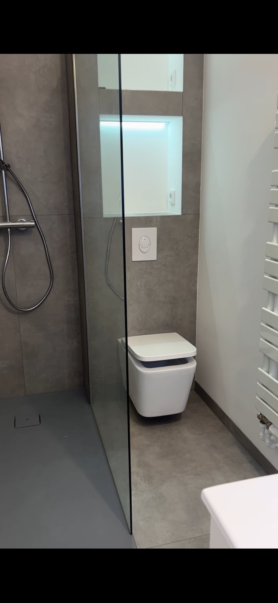 Nowoczesna łazienka z kabiną prysznicową typu walk-in, podwieszaną toaletą z przyciskiem w kształcie uśmiechu i grzejnikiem drabinkowym w kolorze białym, szare płytki na ścianach i podłodze.