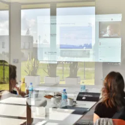 Spotkanie online z doradcą podatkowym na ekranie projektora w sali konferencyjnej, widoczne osoby, butelki wody i ciasteczka na stole.