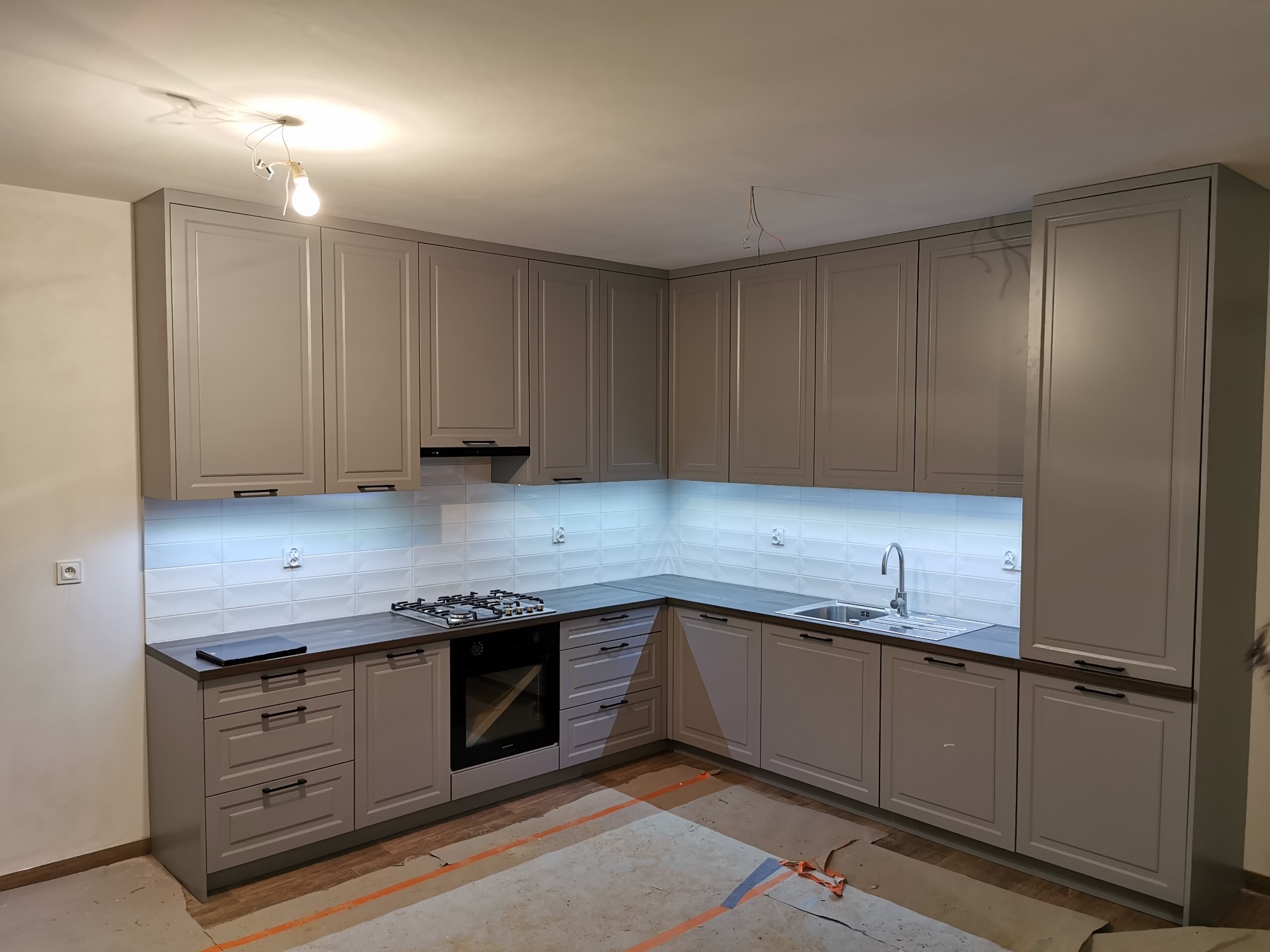 Szara, narożna zabudowa kuchenna z ciemnym blatem i oświetleniem LED pod szafkami wiszącymi, widoczne niedokończone prace wykończeniowe podłogi.
