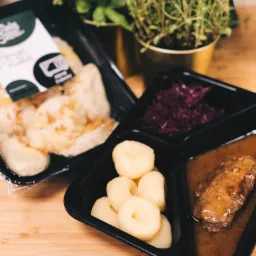 Gotowe danie w czarnym opakowaniu na wynos: kluski śląskie, modra kapusta i mięso w sosie, obok pierogi w folii i zioła w złotych doniczkach.