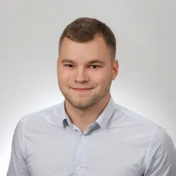 Portret młodego mężczyzny w jasnej koszuli na jednolitym szarym tle, uśmiechający się, z krótkimi włosami i lekkim zarostem.