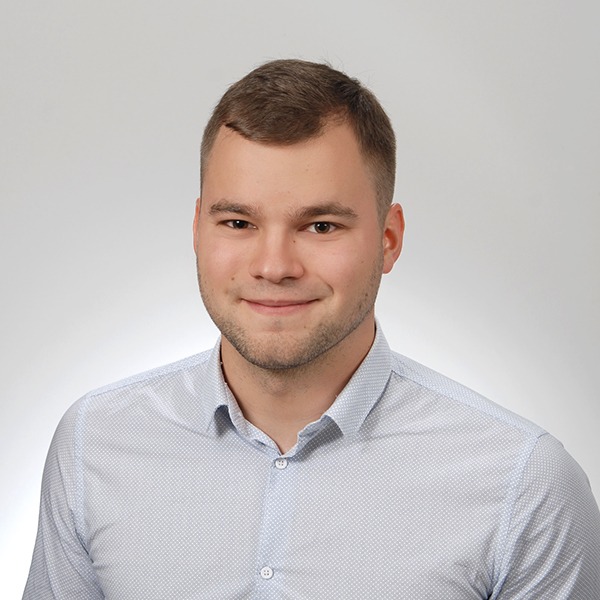 Portret młodego mężczyzny w jasnej koszuli na jednolitym szarym tle, uśmiechający się, z krótkimi włosami i lekkim zarostem.