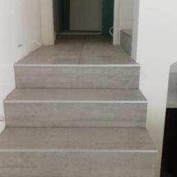 Walerczak Marcin - Schody wyłożone szarymi płytkami ceramicznymi z metalowymi listwami krawędziowymi, prowadzące do pomieszczenia z drzwiami w tle.