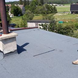 Walerczak Marcin - Pracownik w rękawicach roboczych chodzi po płaskim dachu pokrytym szarą papą, widoczny komin z szarym betonowym obłożeniem, w tle zabudowania wiejskie.