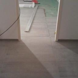 Walerczak Marcin - Ułożone szare płytki podłogowe w przejściu między pomieszczeniami, widoczne materiały budowlane i przewody w tle.