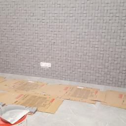 Walerczak Marcin - Ściana wykończona szarymi, trójwymiarowymi panelami kwadratowymi; podłoga zabezpieczona kartonem, widoczne narzędzia i pojemnik z farbą.