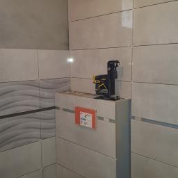 Walerczak Marcin - Ściana łazienki w trakcie układania płytek, widoczny laser krzyżowy Stanley, poziomica, rury kanalizacyjne i płytki z efektem falowania.