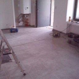 Walerczak Marcin - Wnętrze w trakcie prac wykończeniowych: świeżo ułożona szara podłoga z płytek, drabina aluminiowa z lewej strony, narzędzia i sprzęt budowlany rozłożone w pomieszczeniu, piec gazowy na ścianie...