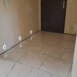 Walerczak Marcin - Ułożone białe płytki podłogowe z szarym marmurkowym wzorem, wbudowane okrągłe lampki w ścianie, szara drzwi w tle, widoczne ślady prac wykończeniowych na ścianach.