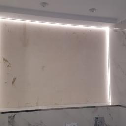 Walerczak Marcin - Ściana w łazience z marmurowymi płytkami, oświetleniem LED w ramce i surową ścianą wnęki z notatkami ołówkiem, widoczny fragment kabiny prysznicowej.