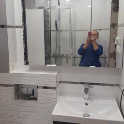 Walerczak Marcin - Wykończona łazienka z białą umywalką na ciemnoszarej szafce, lustro z szafką nad umywalką, widoczna kabina prysznicowa w odbiciu lustra, osoba robiąca zdjęcie telefonem.