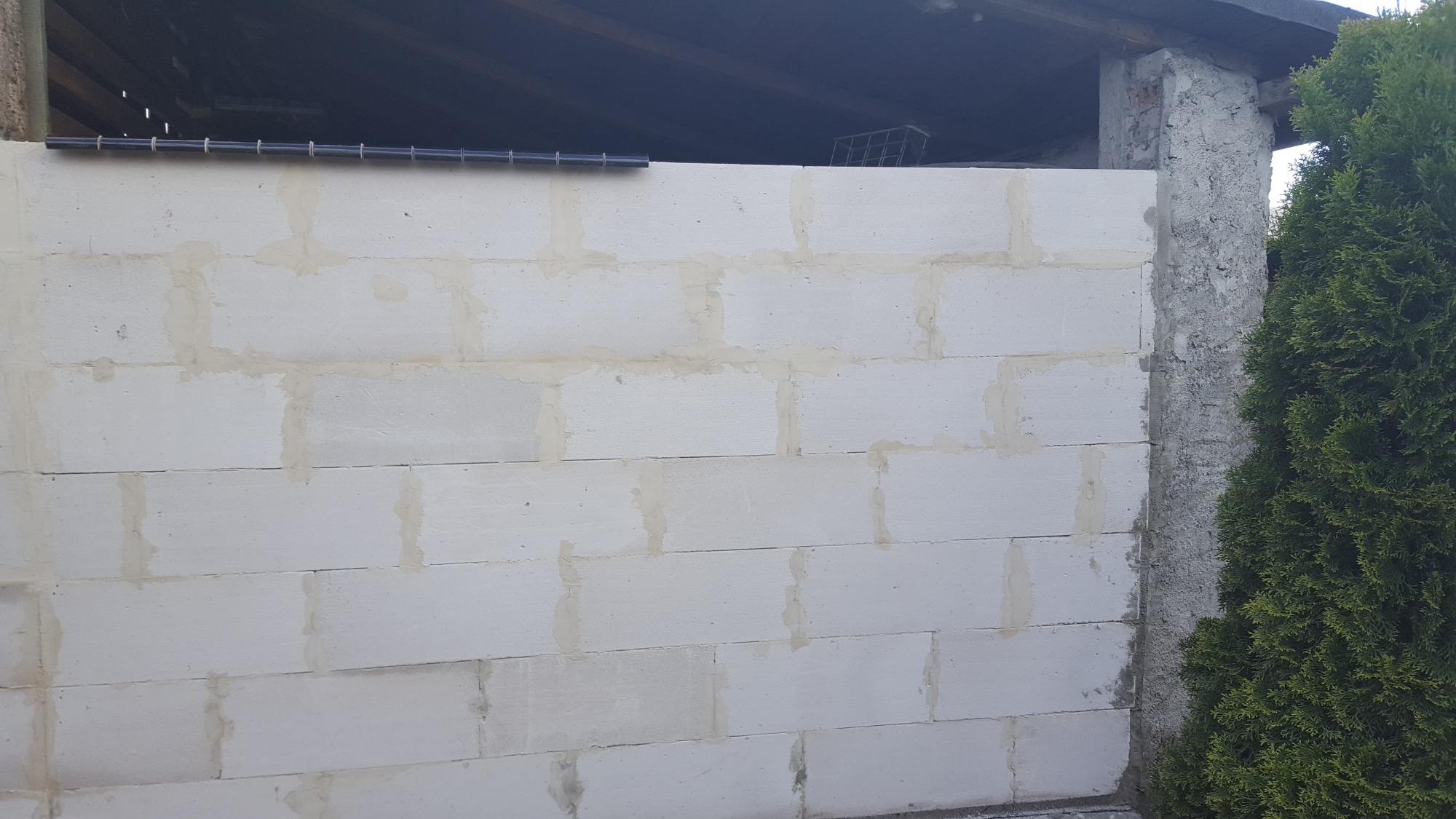 Ściana z białych bloczków betonowych, częściowo wykończona, z widocznymi spoinami i fragmentem konstrukcji dachu w tle, obok rosnący krzew.