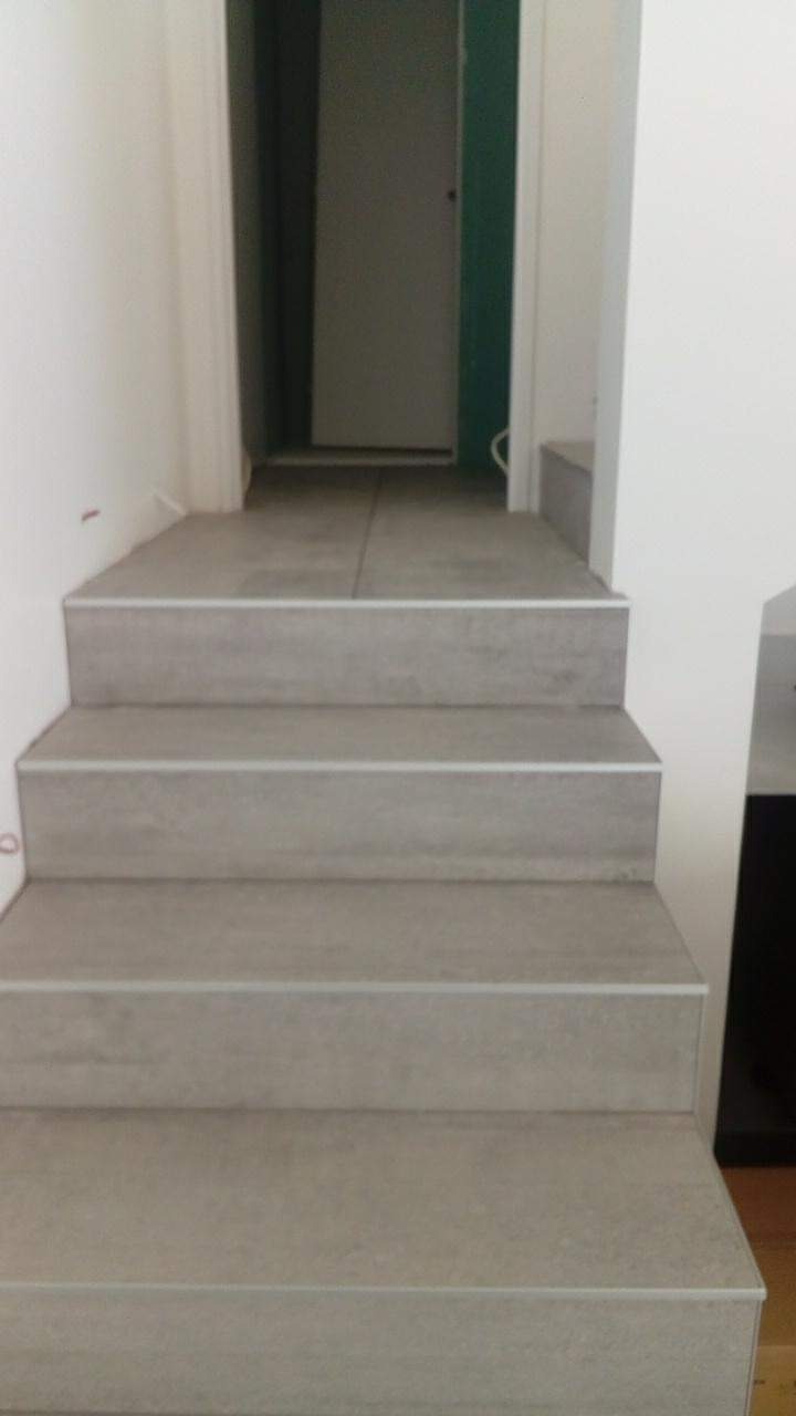 Schody wyłożone szarymi płytkami ceramicznymi z metalowymi listwami krawędziowymi, prowadzące do pomieszczenia z drzwiami w tle.