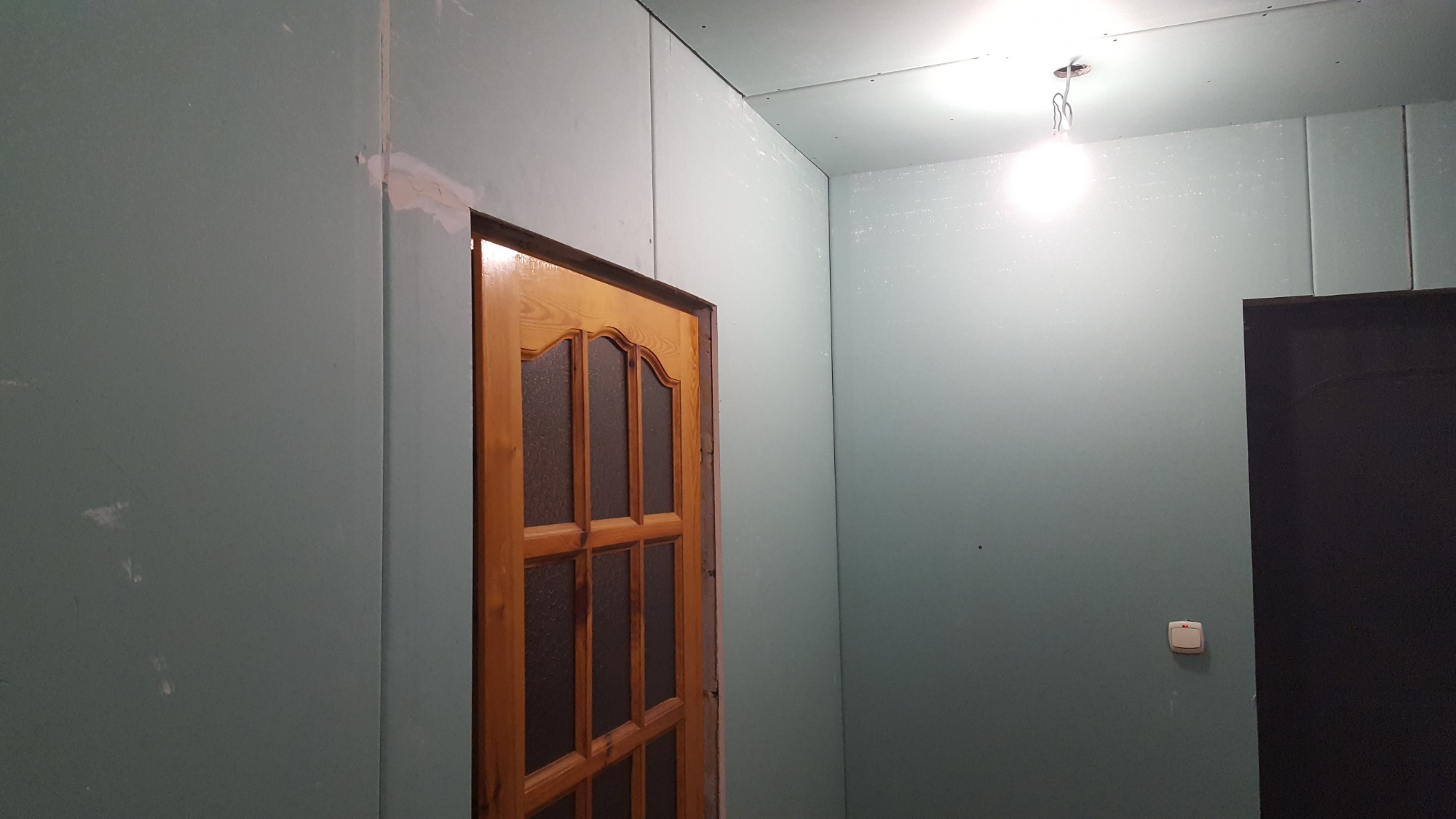 Ściany pokryte płytami kartonowo-gipsowymi w trakcie montażu, widoczne łączenia wymagające szpachlowania, ościeżnica drewniana drzwi wewnętrznych, surowa żarówka na suficie.