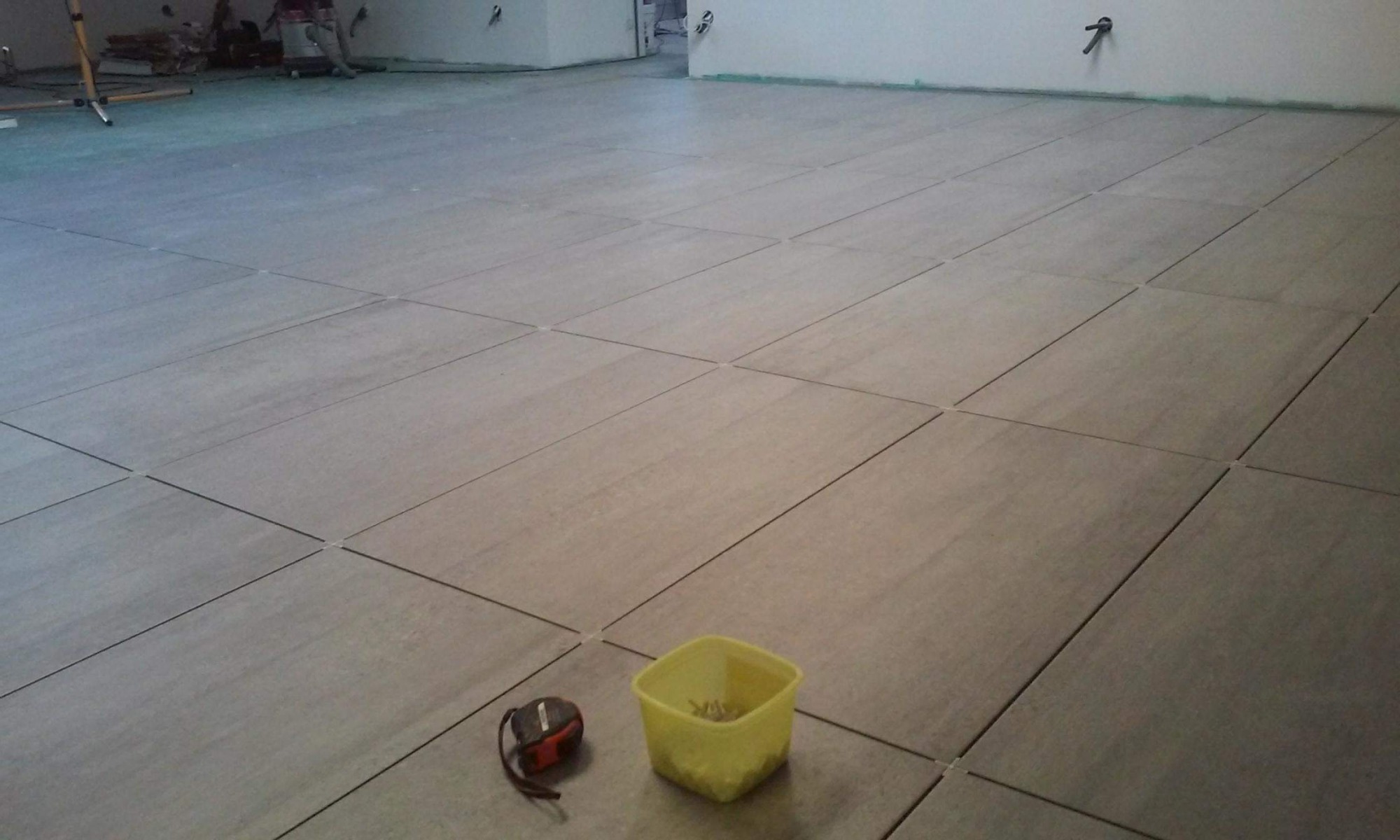 Ułożone płytki podłogowe w dużym pomieszczeniu, widoczne krzyżyki dystansowe, miarka i żółty pojemnik z narzędziami, w tle pracownik.