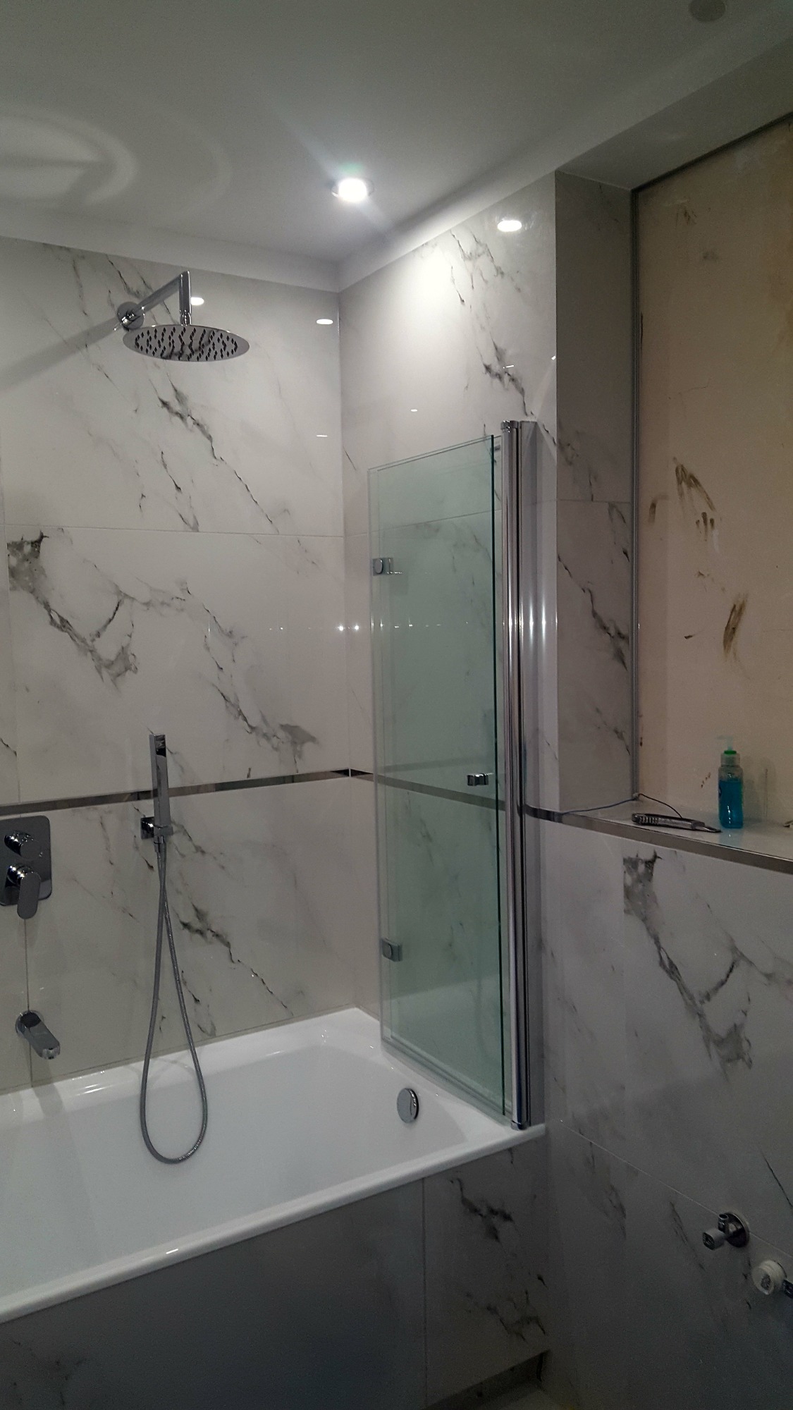 Nowoczesna łazienka z białą wanną, szklanym parawanem nawannowym i marmurowymi płytkami ściennymi z szarymi żyłami, oświetlona punktowymi lampami sufitowymi.