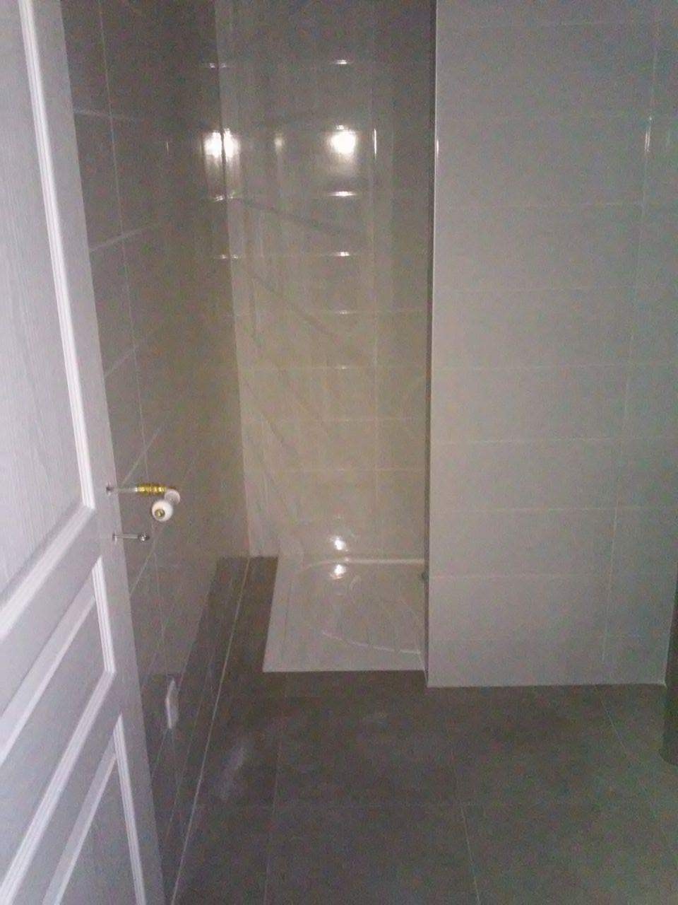 Wykończona kabina prysznicowa z szarymi płytkami, widoczny fragment białych drzwi z chromowaną klamką.