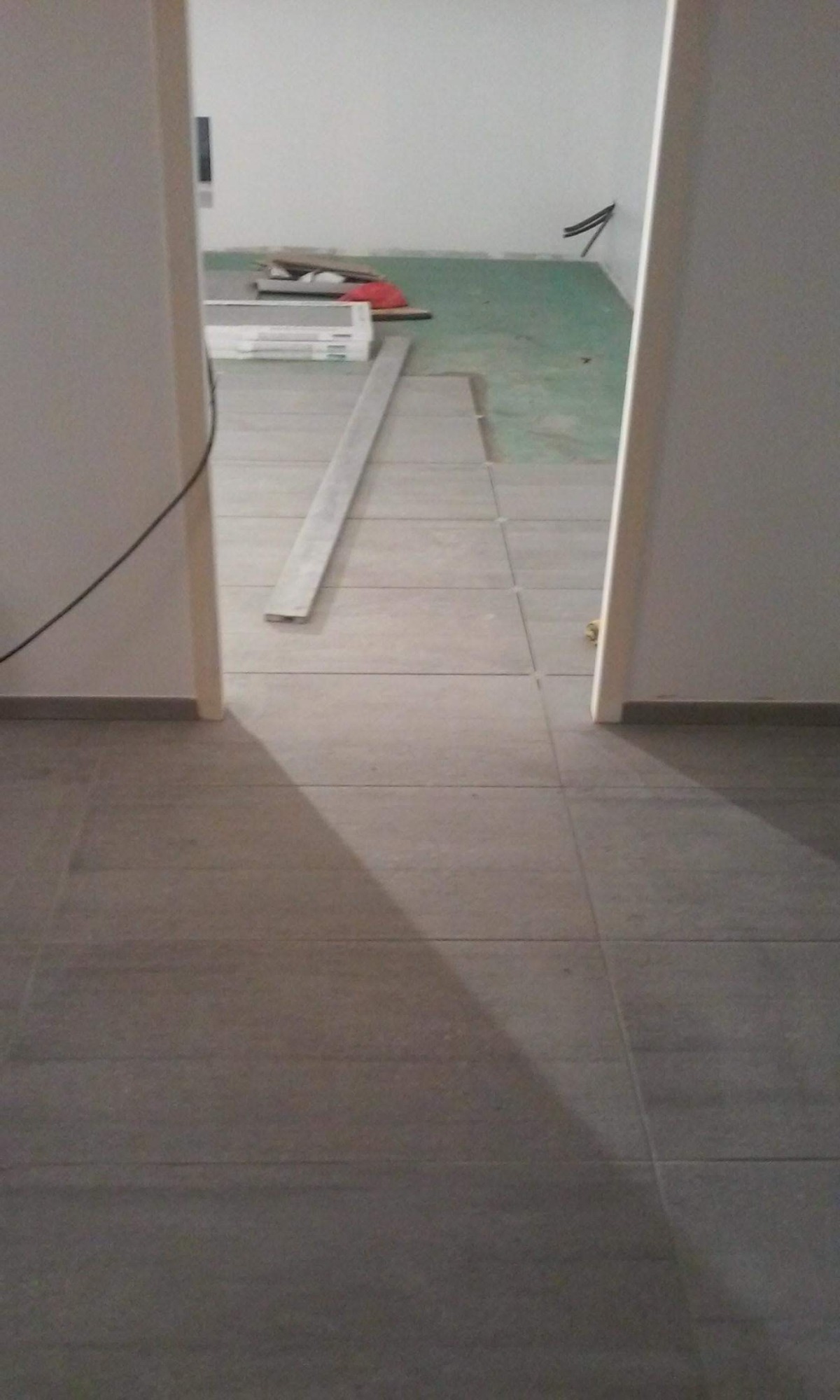 Ułożone szare płytki podłogowe w przejściu między pomieszczeniami, widoczne materiały budowlane i przewody w tle.