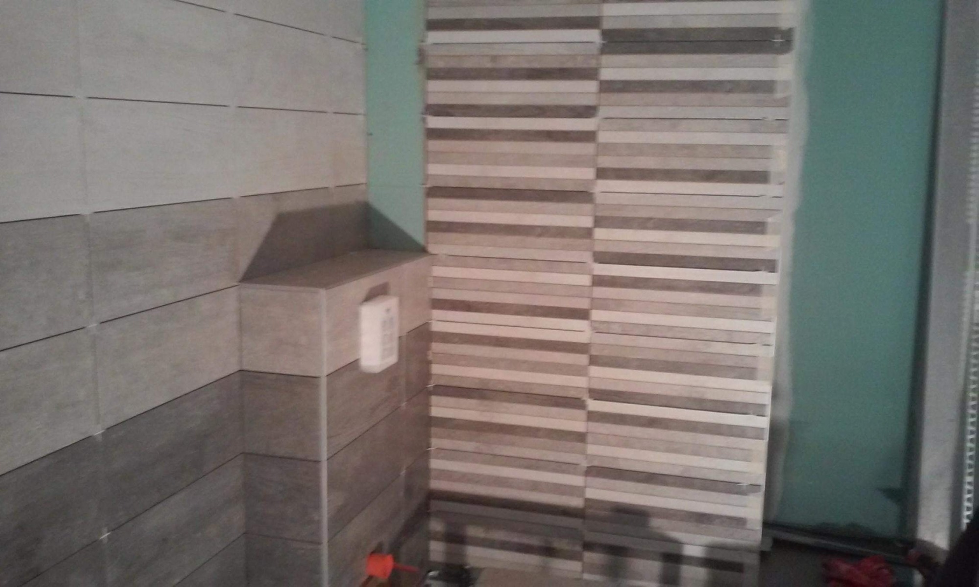 Ściana łazienki z częściowo ułożonymi płytkami ceramicznymi w odcieniach szarości i beżu, z widoczną instalacją podtynkową i stosem płytek przygotowanych do montażu na tle surowej, zielonej ściany.