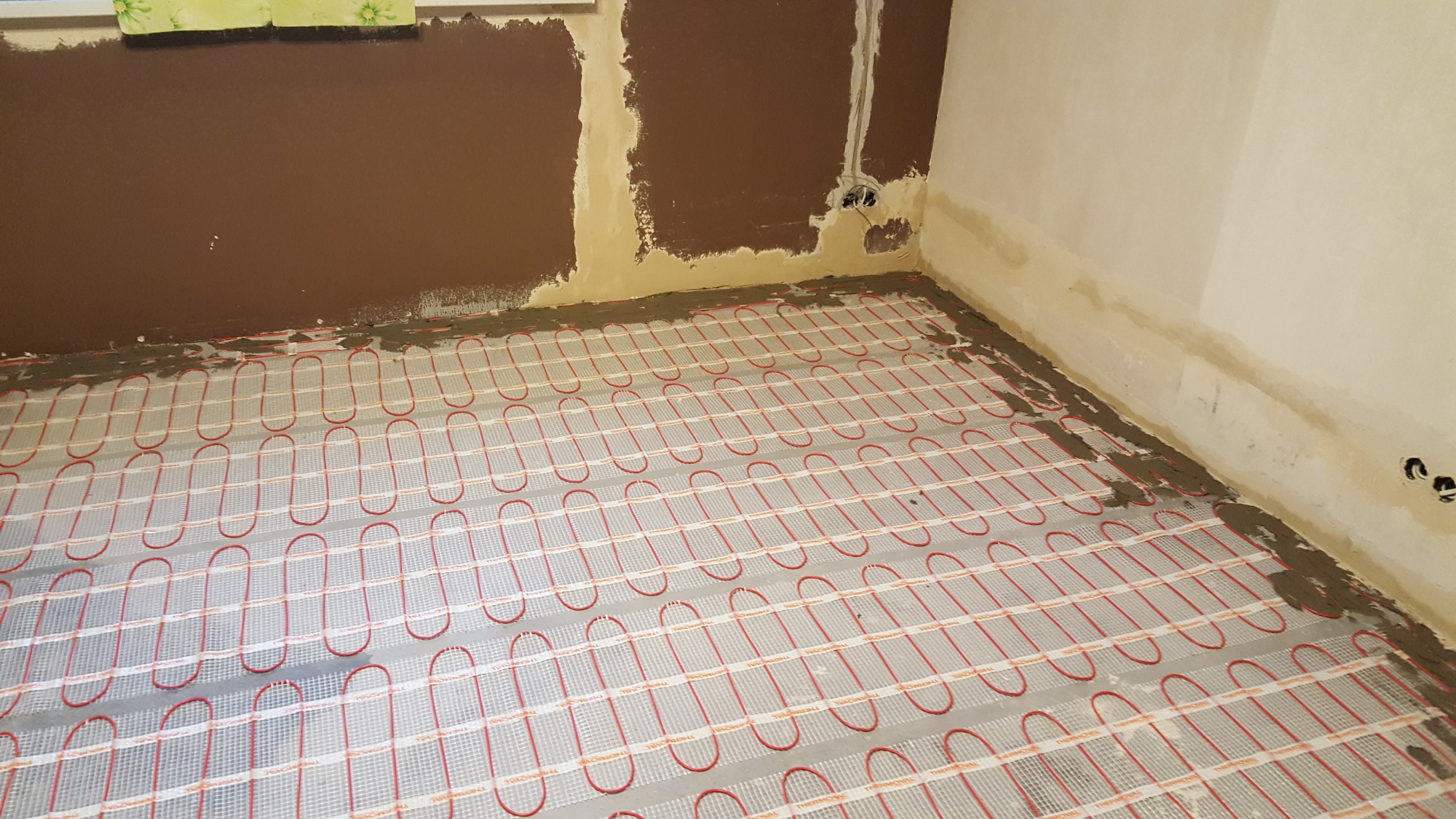 Rozłożona mata grzewcza pod podłogę w trakcie prac przygotowawczych, widoczne ślady zaprawy na ścianach i przewodach.