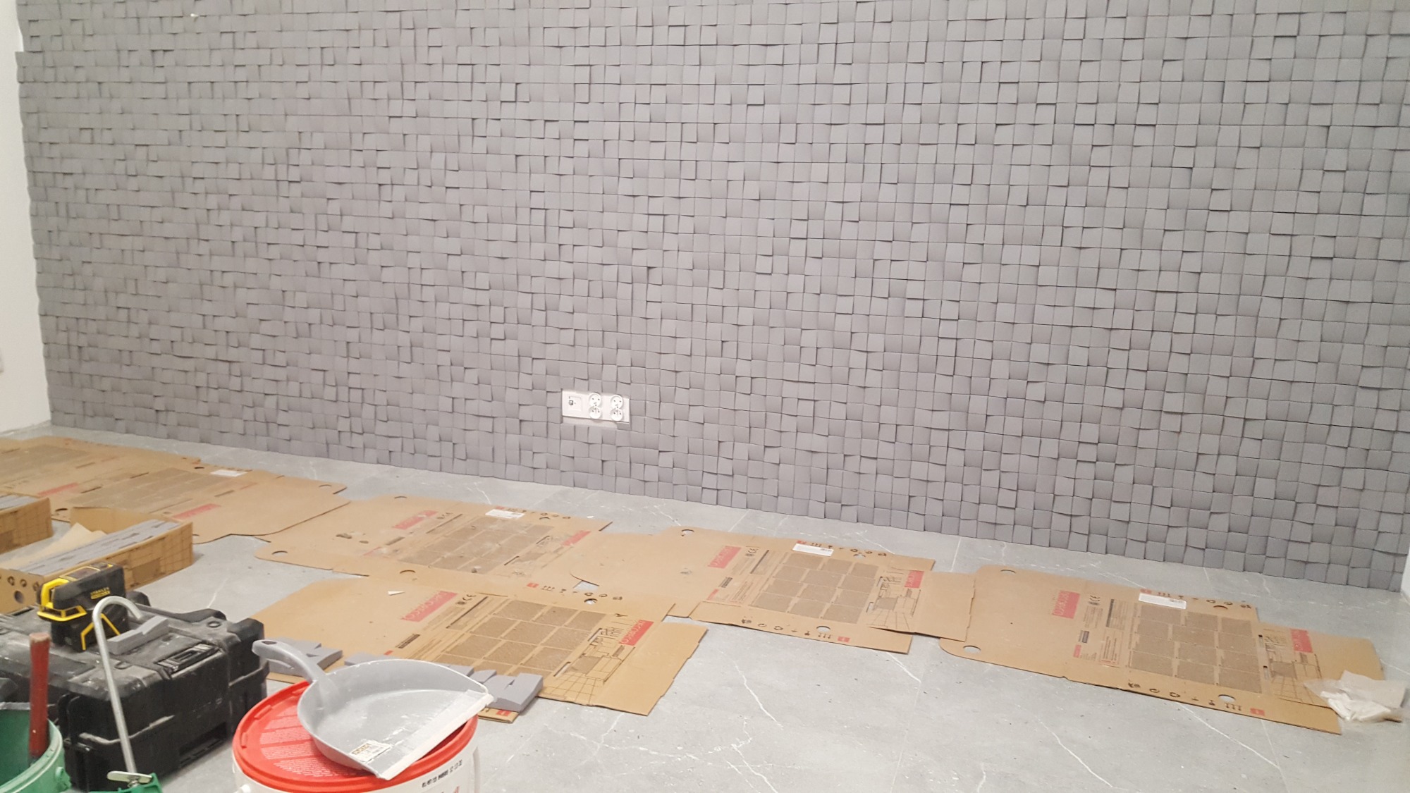 Ściana wykończona szarymi, trójwymiarowymi panelami kwadratowymi; podłoga zabezpieczona kartonem, widoczne narzędzia i pojemnik z farbą.