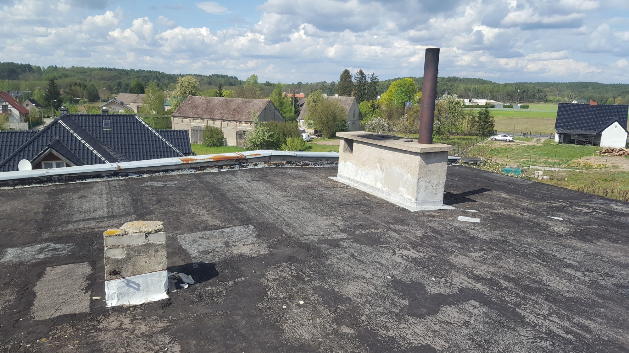 Niedawno naprawiony, płaski dach pokryty papą z widocznymi łatami, dwa kominy cementowe z metalowymi rurami, w tle zabudowania wiejskie i drzewa.