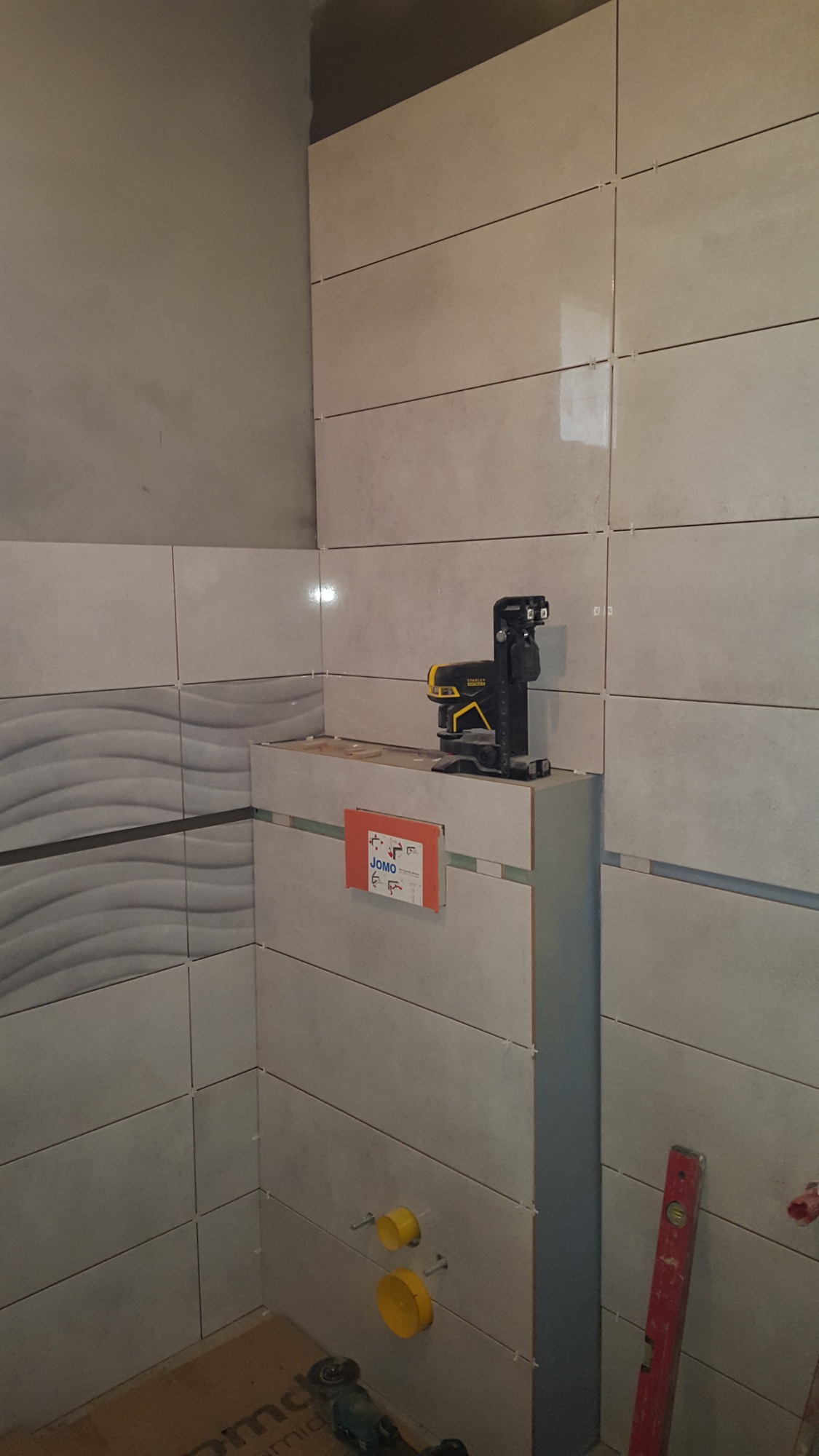 Ściana łazienki w trakcie układania płytek, widoczny laser krzyżowy Stanley, poziomica, rury kanalizacyjne i płytki z efektem falowania.