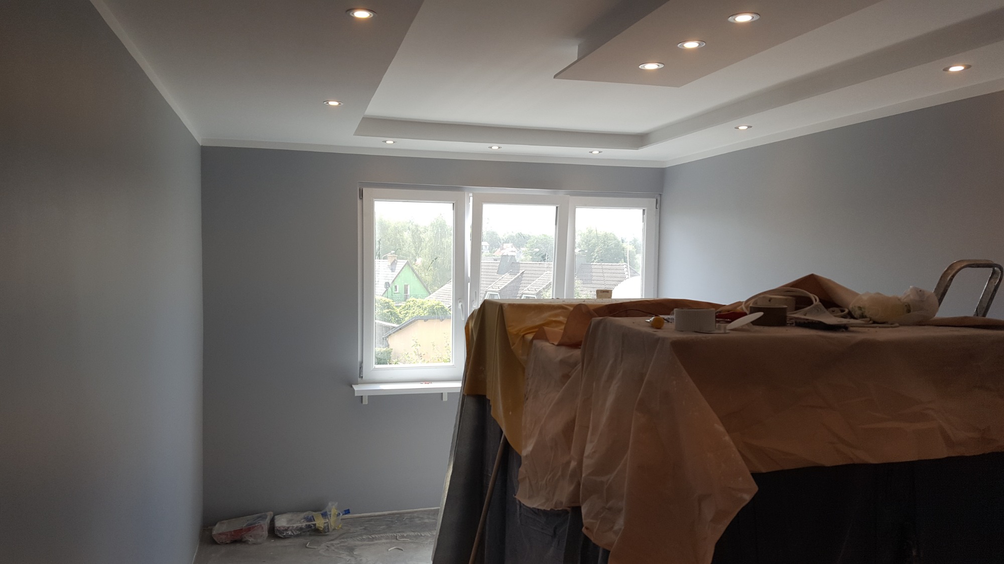 Świeżo pomalowane na szaro ściany pomieszczenia z zamontowanym oświetleniem punktowym w suficie podwieszanym, widok zza osłoniętych folią mebli i narzędzi malarskich.