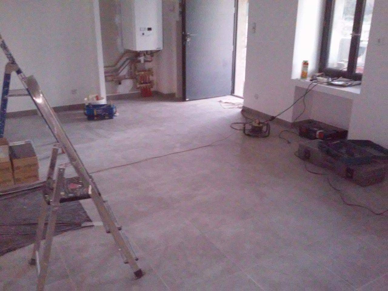 Wnętrze w trakcie prac wykończeniowych: świeżo ułożona szara podłoga z płytek, drabina aluminiowa z lewej strony, narzędzia i sprzęt budowlany rozłożone w pomieszczeniu, piec gazowy na ścianie...