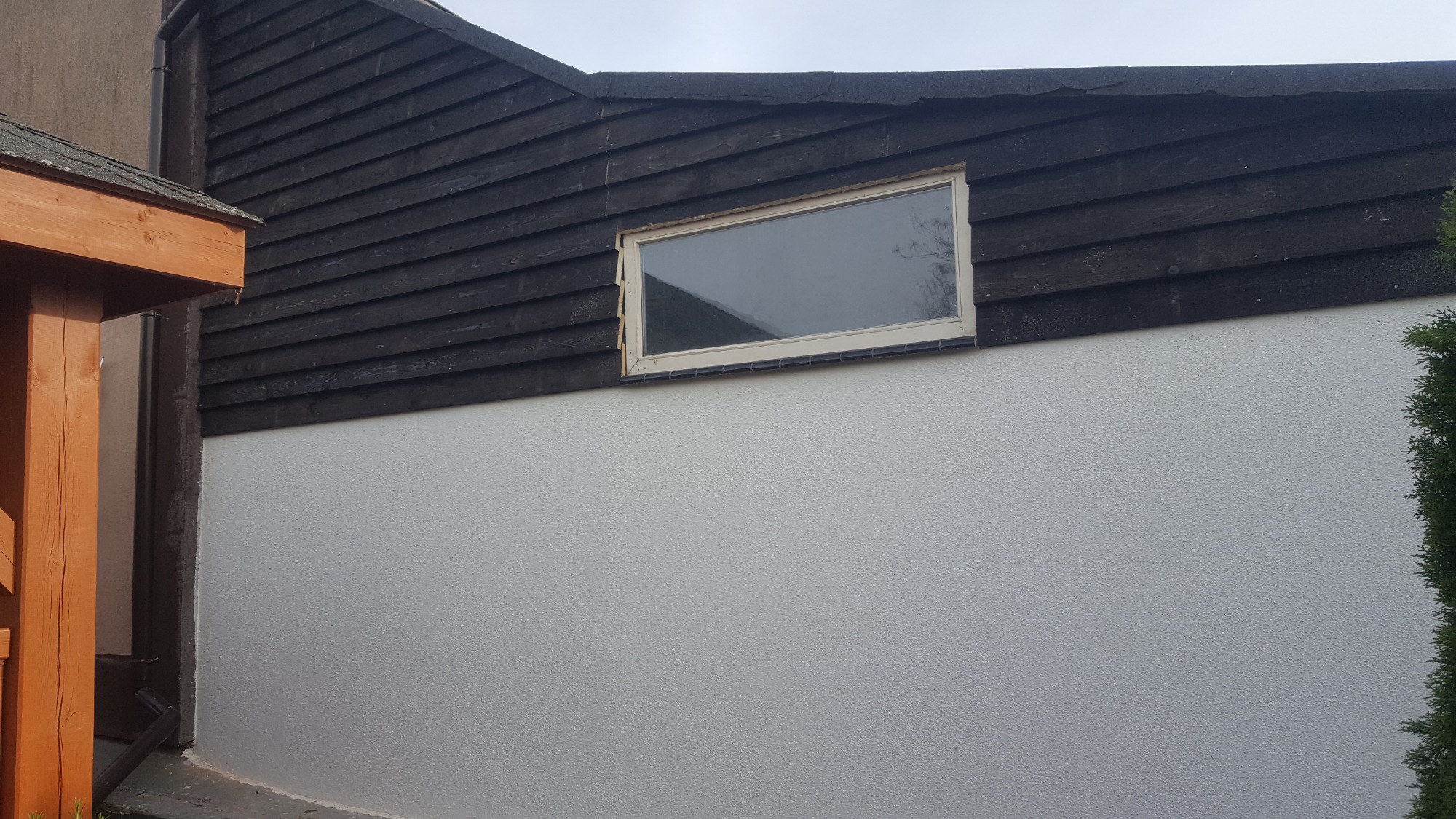 Ściana budynku z oknem, pomalowana na biało w dolnej części i wykończona ciemnym drewnem w górnej części, widoczne nierówności faktury tynku.