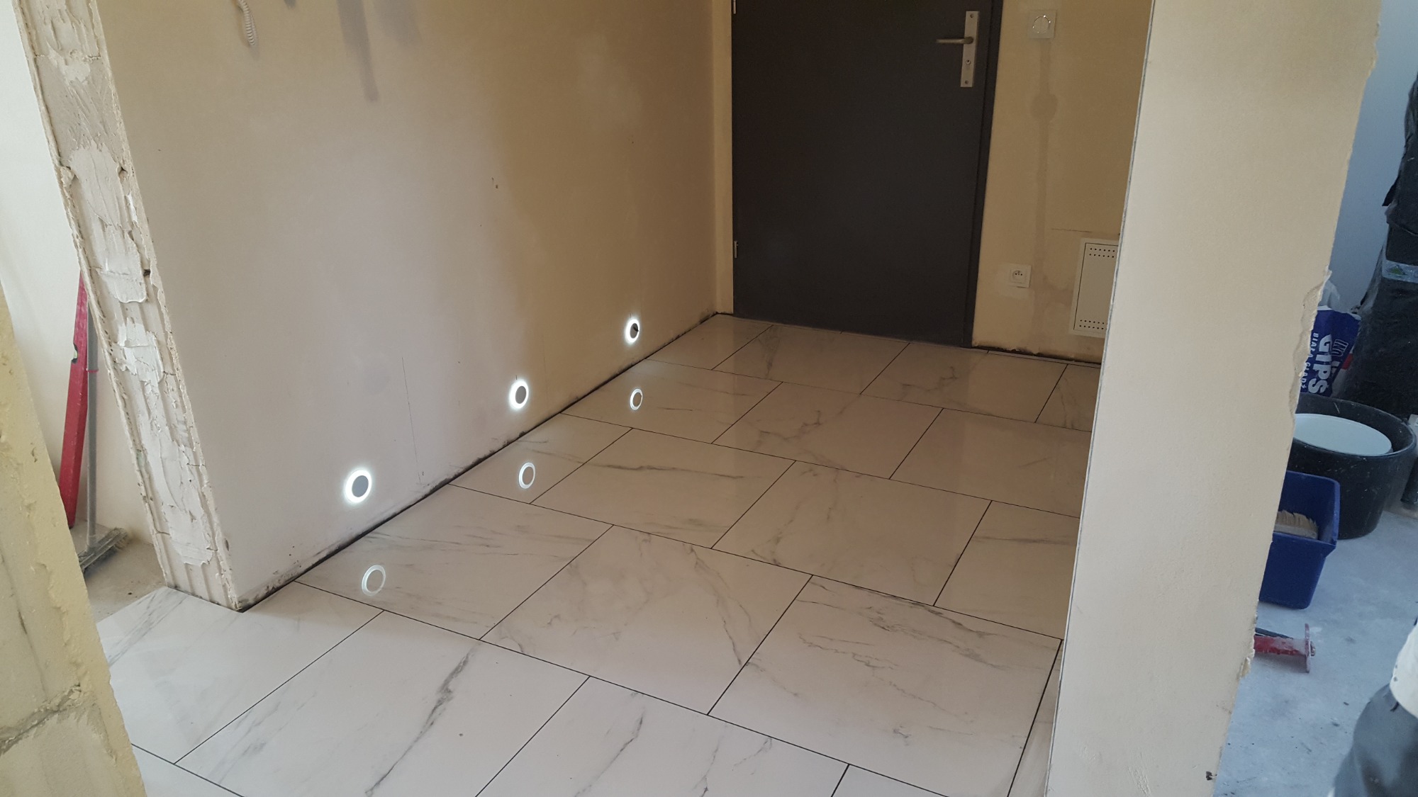 Ułożone białe płytki podłogowe z szarym marmurkowym wzorem, wbudowane okrągłe lampki w ścianie, szara drzwi w tle, widoczne ślady prac wykończeniowych na ścianach.