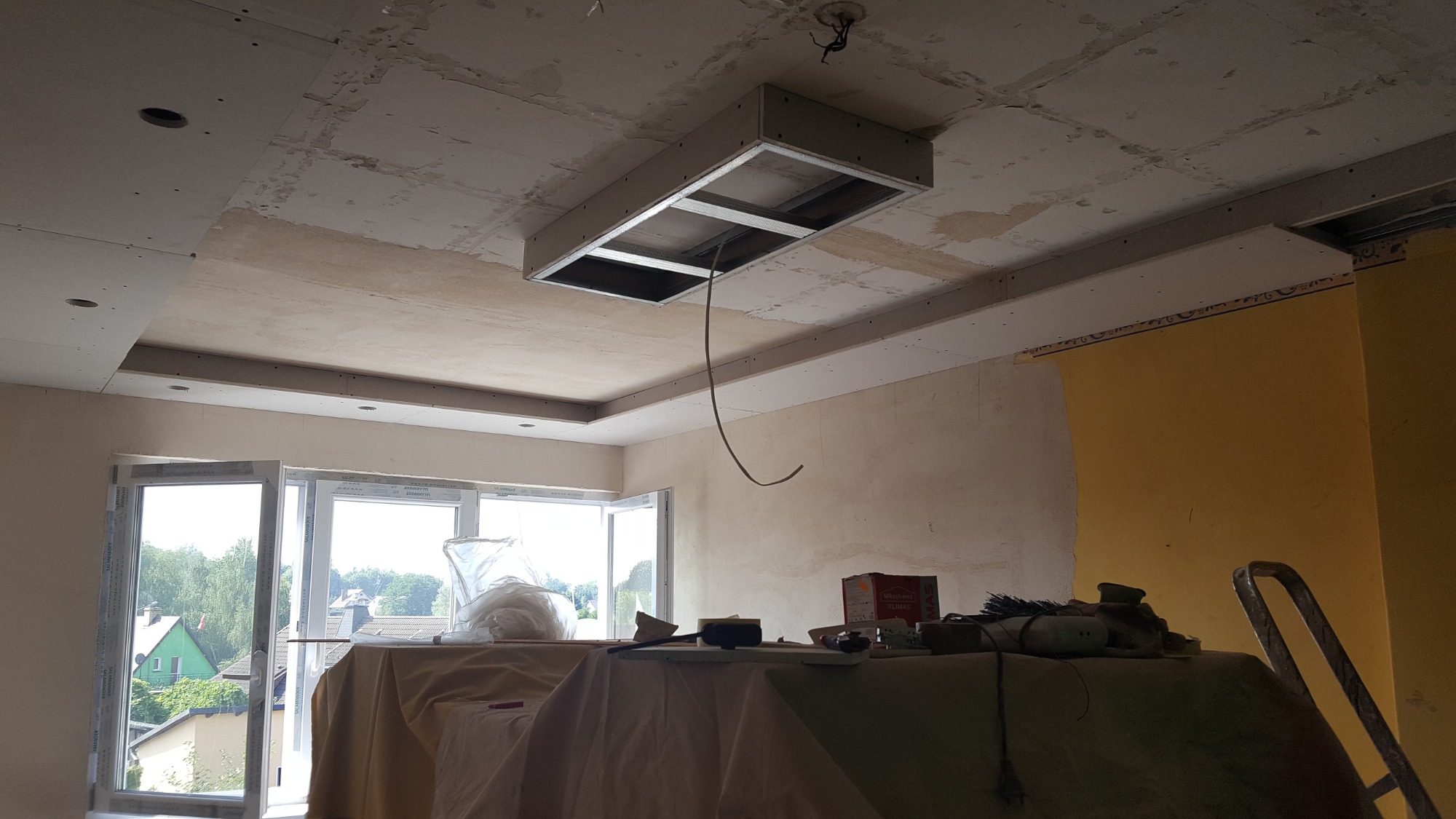 Sufit w trakcie montażu z widoczną konstrukcją podwieszaną, częściowo pokryty płytami kartonowo-gipsowymi, z otwartym oknem w tle i narzędziami na zakrytym stole.