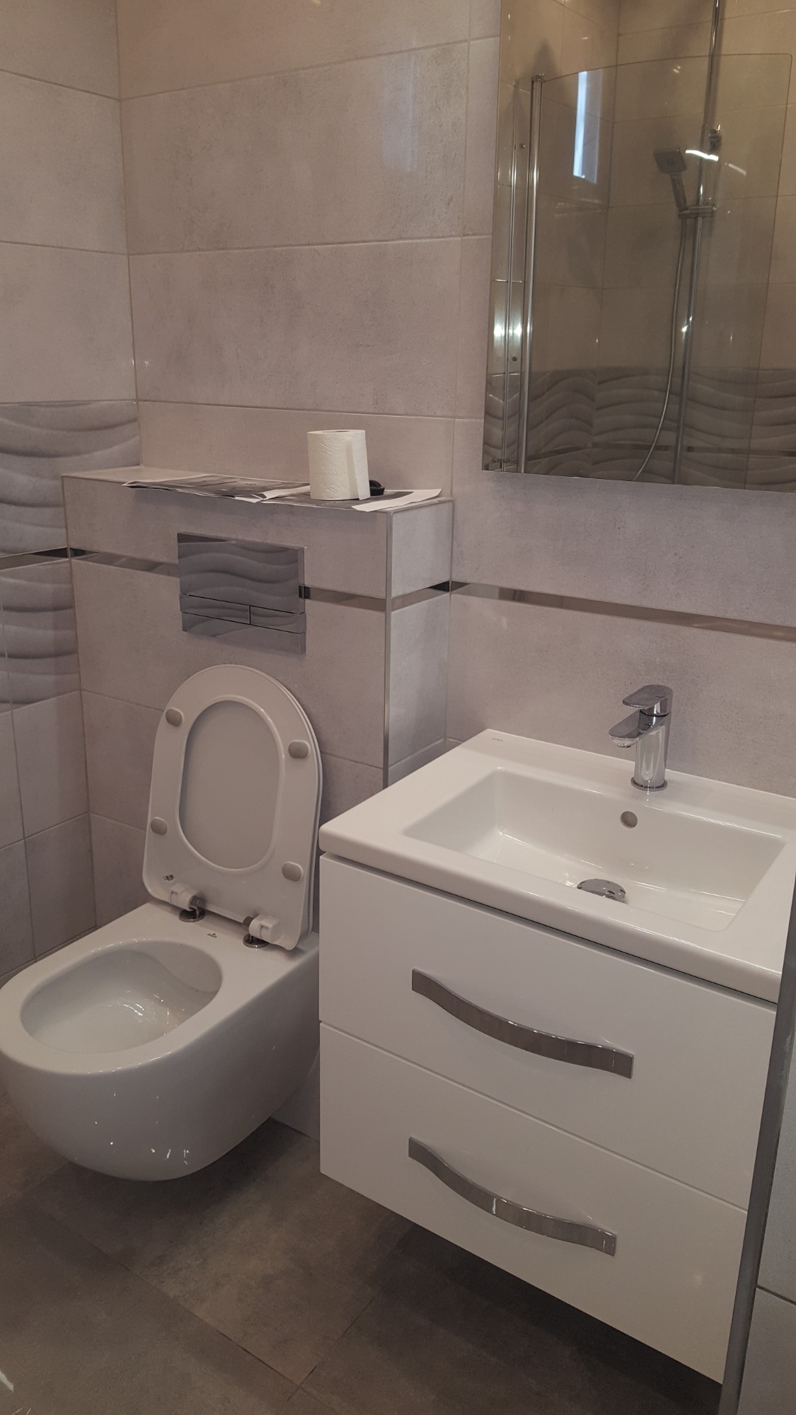 Nowoczesna łazienka z podwieszaną toaletą, umywalką z szafką i szarymi płytkami imitującymi kamień, akcentowana poziomymi metalowymi listwami.