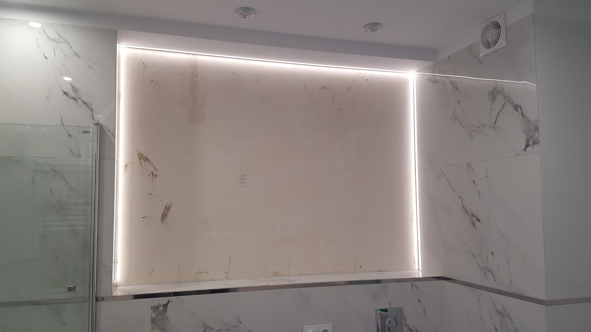 Ściana w łazience z marmurowymi płytkami, oświetleniem LED w ramce i surową ścianą wnęki z notatkami ołówkiem, widoczny fragment kabiny prysznicowej.