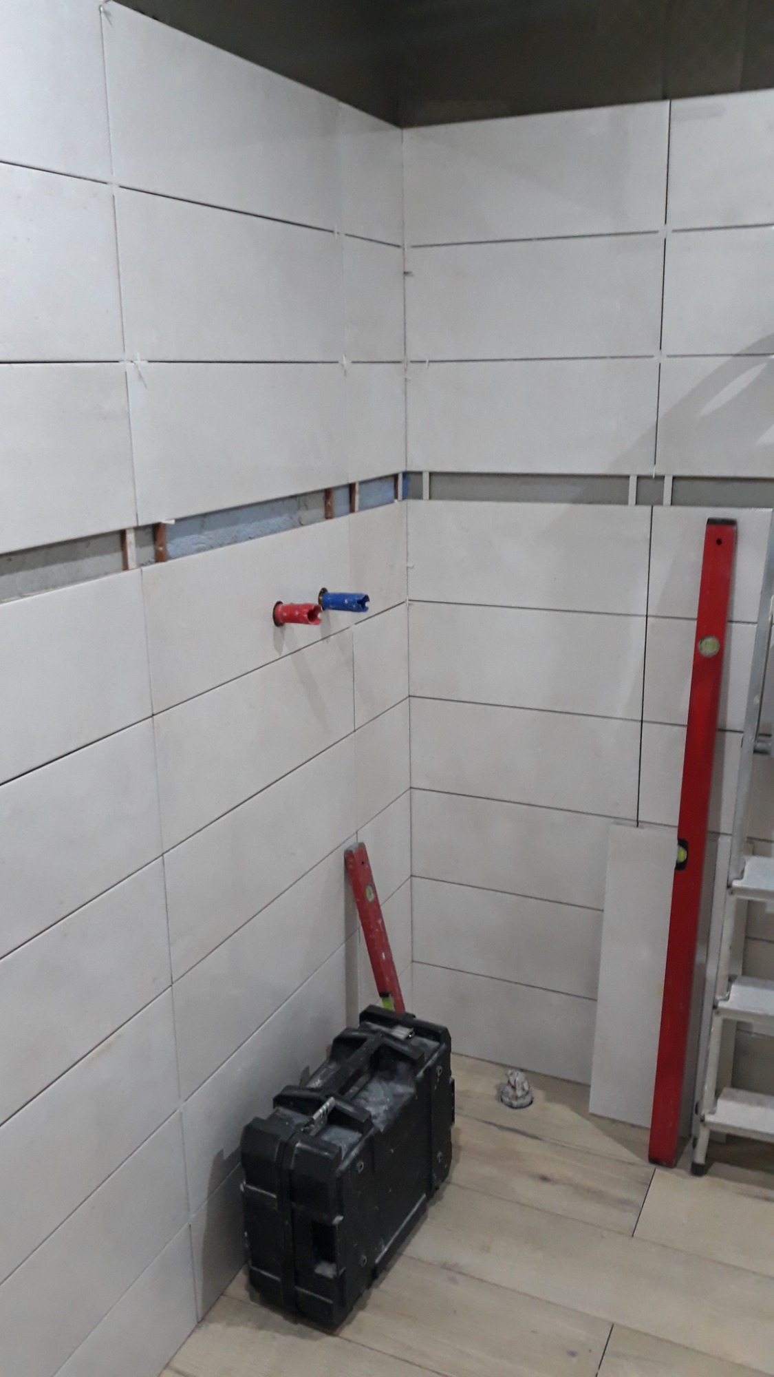 Białe, poziome płytki częściowo ułożone na ścianie łazienki, widoczne czerwone i niebieskie rurki instalacji wodnej, czarna skrzynka narzędziowa na drewnopodobnej podłodze, poziomica i drabina w tle.