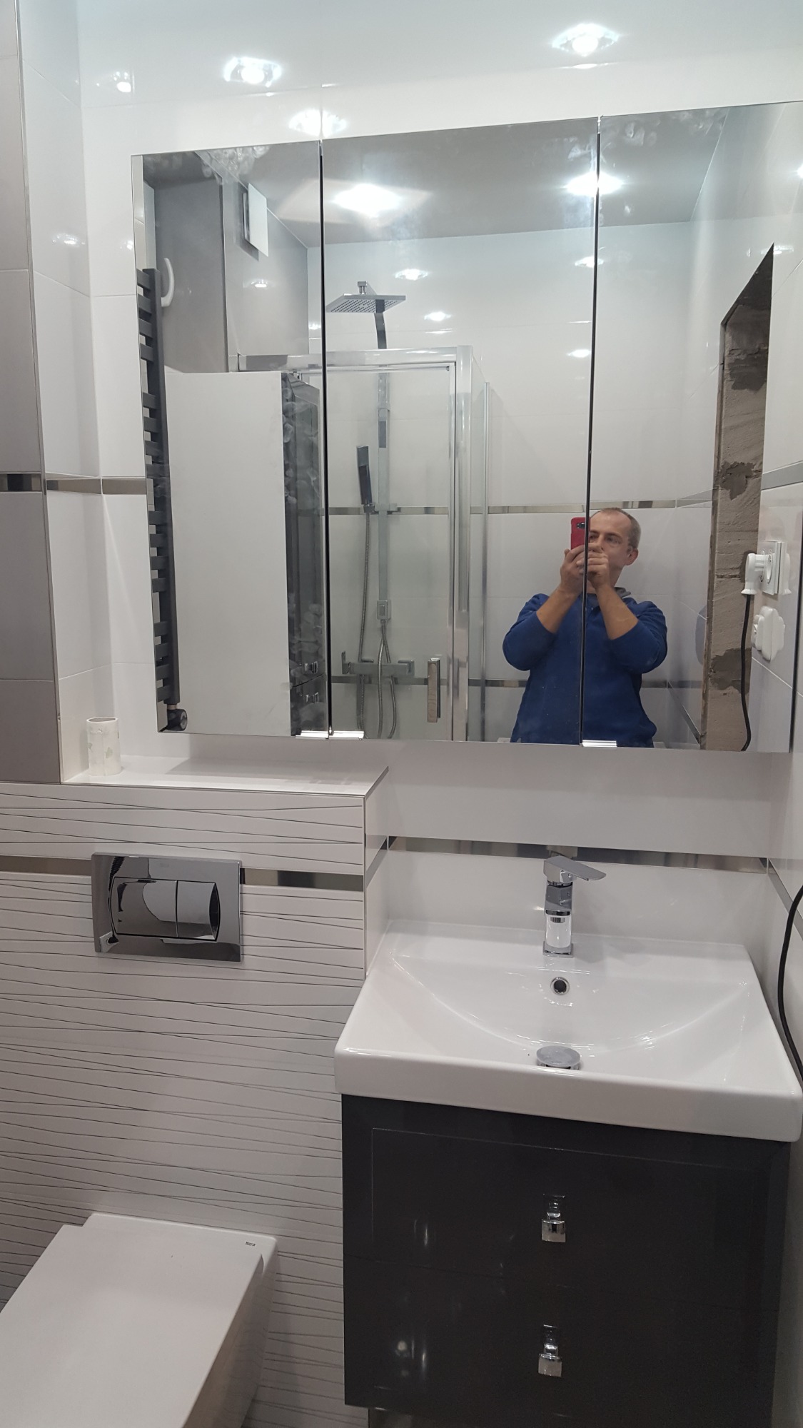 Wykończona łazienka z białą umywalką na ciemnoszarej szafce, lustro z szafką nad umywalką, widoczna kabina prysznicowa w odbiciu lustra, osoba robiąca zdjęcie telefonem.