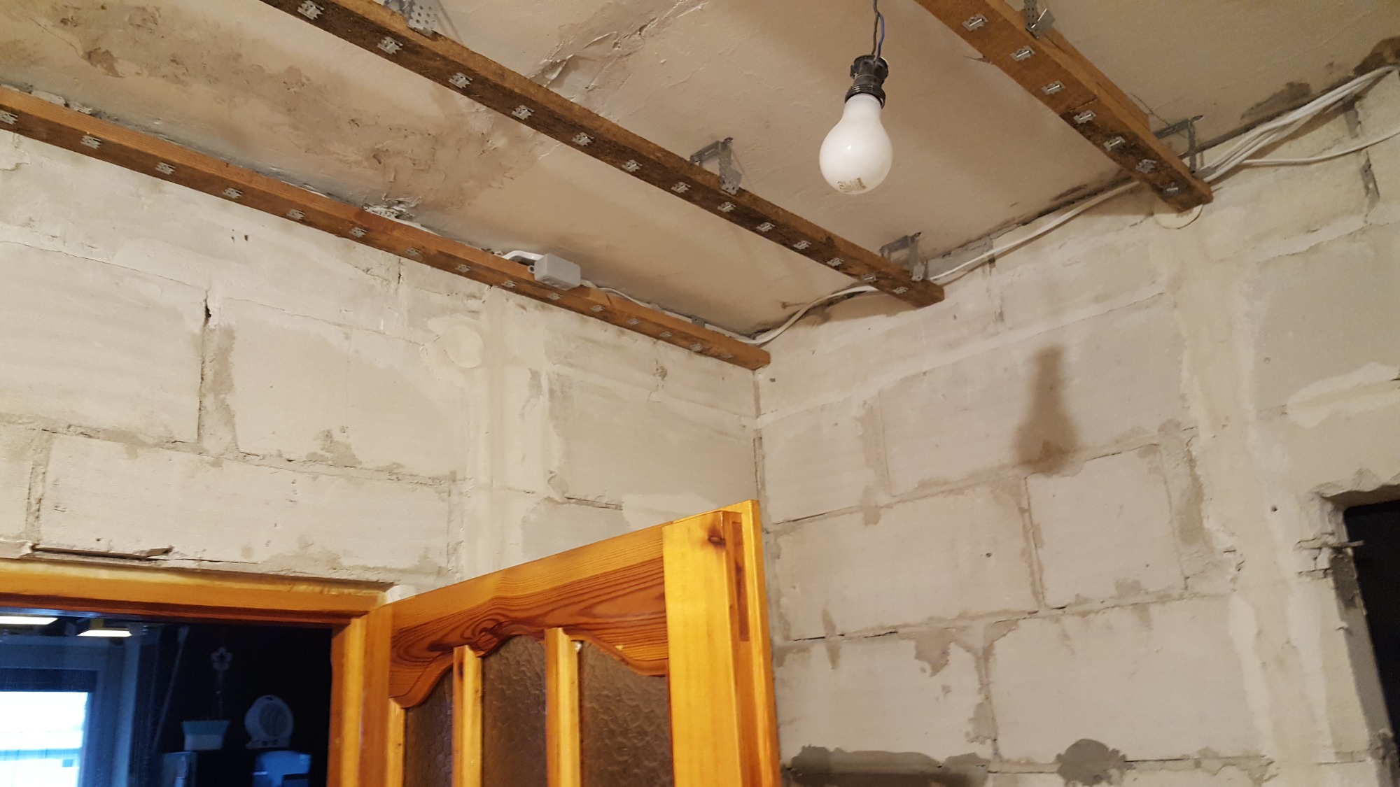 Surowy stan wnętrza: ściany z betonu komórkowego, drewniane stelaże pod sufitem z zamontowanymi punktami świetlnymi, wisząca żarówka, widoczne przewody elektryczne i fragment drewnianych drzwi.